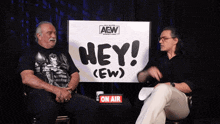 two men sitting in front of a sign that says hey