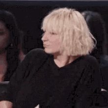 a woman with blonde hair and bangs is sitting in a crowd of people and making a funny face .