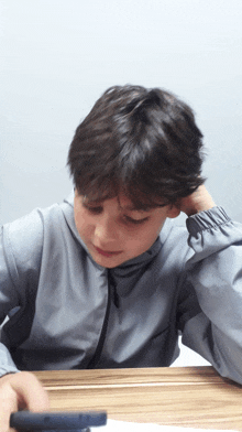 a boy in a grey jacket sits at a table with his hand on his head