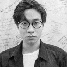 a man wearing glasses stands in front of a wall that has a lot of writing on it