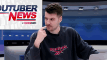 a man wearing a reebok sweatshirt is sitting in front of a pop buzz logo