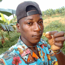 a young man wearing a hat and a hawaiian shirt holds a piece of food in his hand