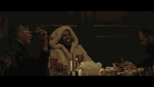 a man wearing a fur coat with a hood is sitting at a table with drinks