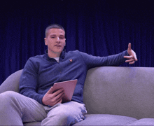 a man is sitting on a couch holding a tablet and wearing a blue jacket with a bird on it