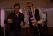 two men are dancing in a room while one holds an american flag