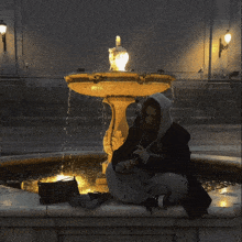 a person in a hoodie sits in front of a fountain at night