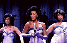 three women are singing into microphones on a stage and one of them is wearing a purple dress