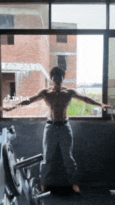 a shirtless man is standing in front of a window with his arms outstretched in a gym .