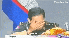 a man is covering his face with his hand while sitting at a table with food in front of him .