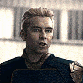 a man with blonde hair and blue eyes is wearing a black jacket and a gold collar .
