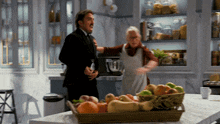 a man in a suit stands next to a woman in an apron in a kitchen with a basket of fruit on the counter