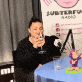 a woman drinking a glass of wine in front of a sign that says subterfuge radio