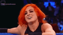 a woman with red hair is smiling in a wrestling ring while leaning on a blue rope .
