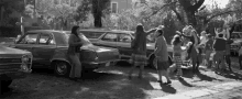 a black and white photo of a group of people dancing in front of a row of cars .