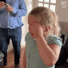 a little girl is covering her face with her hand while a man holds a cell phone in the background .