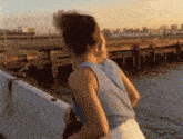 a woman in a blue tank top stands in front of a body of water at sunset