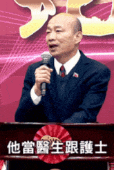 a man in a suit stands at a podium holding a microphone with chinese writing behind him