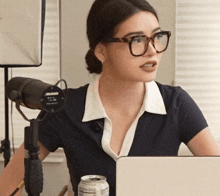 a woman wearing glasses sits in front of a microphone with a can of caffeine in front of her