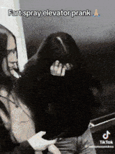 a woman covering her face with her hand while another woman looks at her phone and says fart spray elevator prank