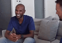a man in a blue shirt sits on a couch smiling