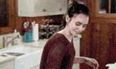 a woman in a red sweater is standing in a kitchen next to a sink and smiling .
