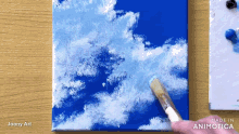a painting of clouds is being painted on a blue canvas by joony art