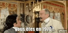 two men are standing next to each other in a room with the words `` vous etes en 1970 '' written on the screen .