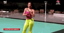 a woman in a pink top and neon yellow pants is dancing in front of a pool .
