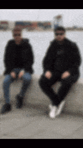 a blurry picture of two men sitting on a wall near the water