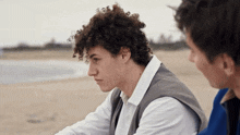a man with curly hair sits on the beach with another man