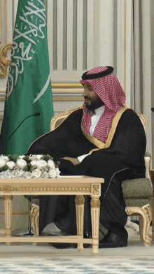 a man in a black robe sits at a table in front of a green flag