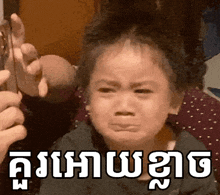 a little girl is crying while being held by a person and the words in a foreign language are visible behind her .