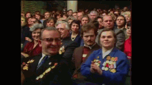 a group of people are sitting in a room and one of them is wearing a blue jacket with medals on it ..