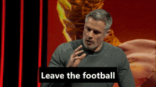 a man in a grey sweater is speaking into a microphone with the words " leave the football " behind him