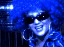 a woman wearing a blue afro wig and sunglasses smiles in front of a disco ball