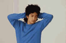 a man with curly hair is wearing a blue sweater and holding his hands behind his head .