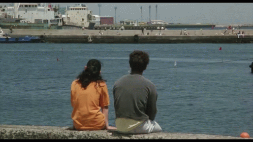 a man and a woman sit on a barrier looking at a shipyard. their backs are to us
