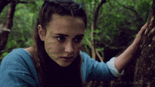 a girl with green leaves on her face is standing next to a tree in the woods