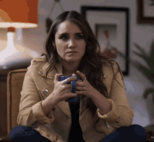 a woman in a tan jacket is sitting in a chair holding a cup of coffee