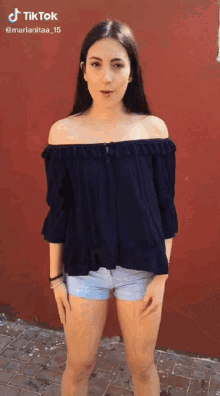 a woman wearing a blue off the shoulder top and shorts stands in front of a red wall ..