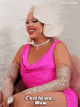 a drag queen wearing a pink dress and gloves is sitting in a chair .