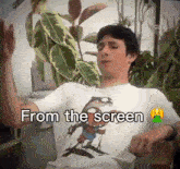 a boy wearing a t-shirt that says from the screen is sitting in front of a plant