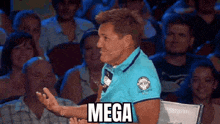 a man in a blue shirt is sitting in front of a microphone in a crowd of people and says mega .