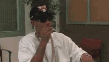 a man wearing sunglasses and a baseball cap is sitting at a table smoking a cigarette .