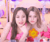 two women are sitting next to each other with balloons in the background and one has a smiley face sticker on her shirt