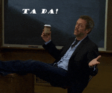 a man in a suit sits in front of a chalkboard that says " ta da "