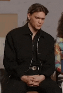 a man in a black jacket is sitting on a chair with his hands folded in front of him .