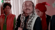 a man with a mustache is standing in front of a group of men wearing turbans and scarves .