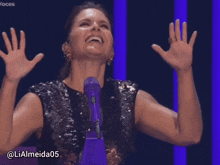 a woman in a black sequined top stands in front of a purple microphone with her hands in the air