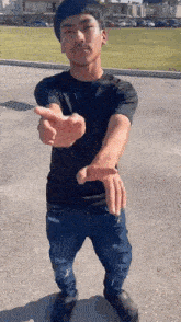 a young man in a black shirt and blue jeans is standing in a parking lot and pointing at the camera .